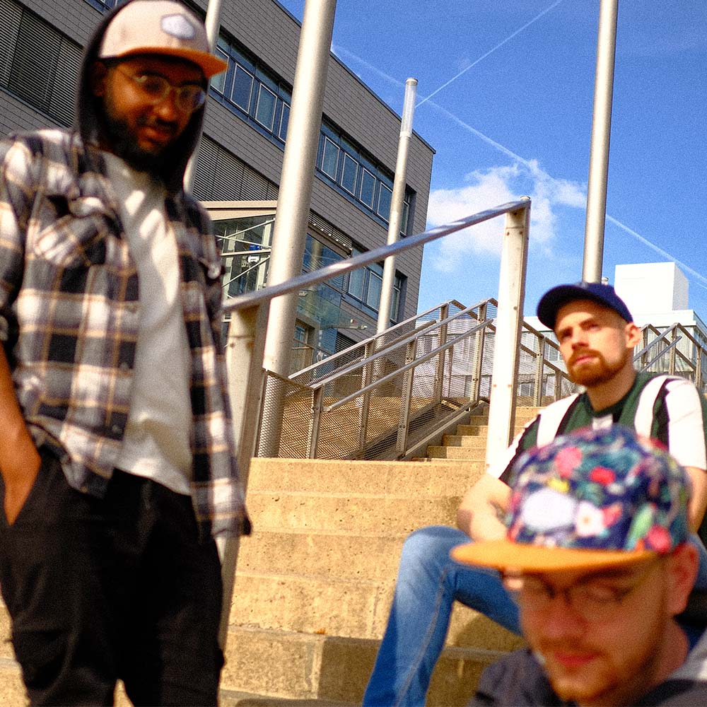 Drei junge Männer sitzen und stehen auf einer Treppe. Alle drei tragen verschiedene extragroße Snapback, Baseball und Fivepanel Caps von BigBrains Co.