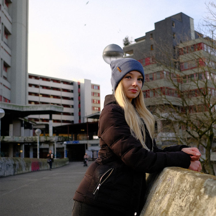 Blonde junge Frau steht vor Betonhochhaus. Sie trägt die extragroße XL Beanie Mütze von BigBrains Co.