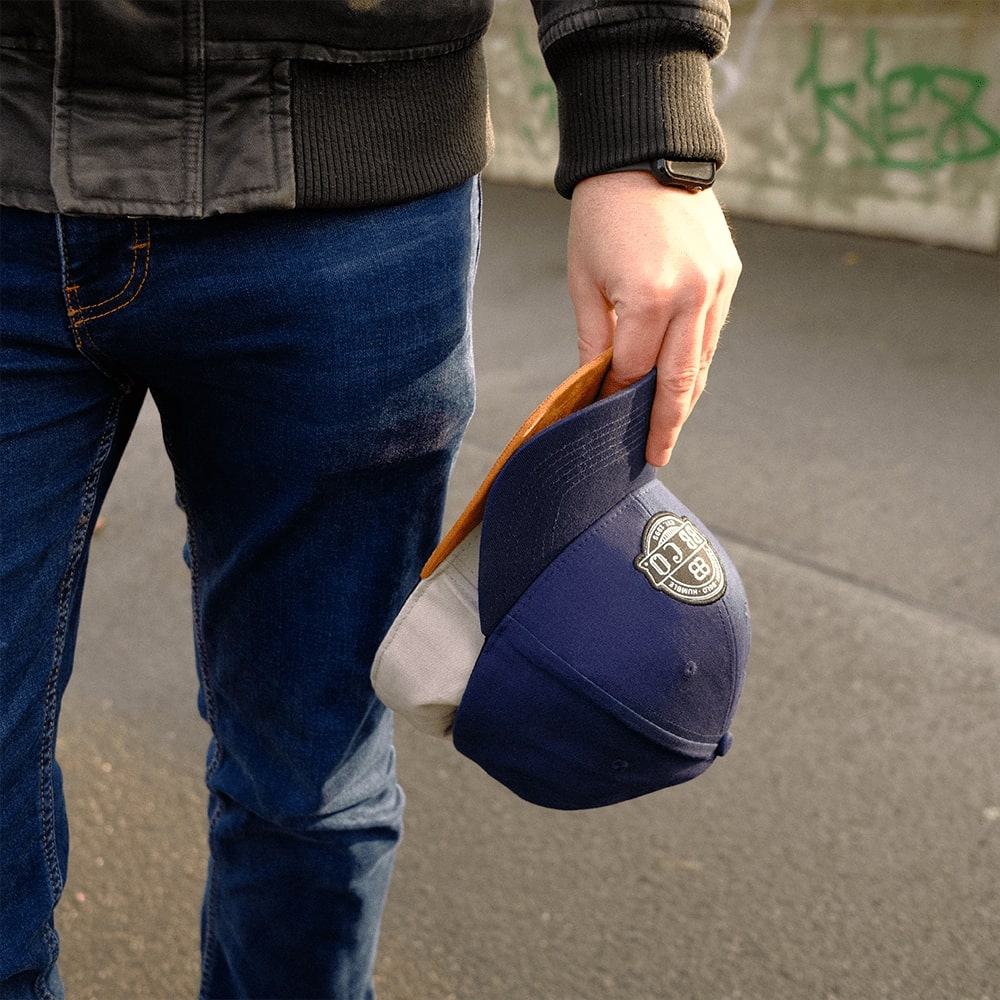 Nahaufnahme auf die Hand eines Mannes der zwei Caps (graue XXL Snapback Cap und blaue oversize Baseball Cap) hält.