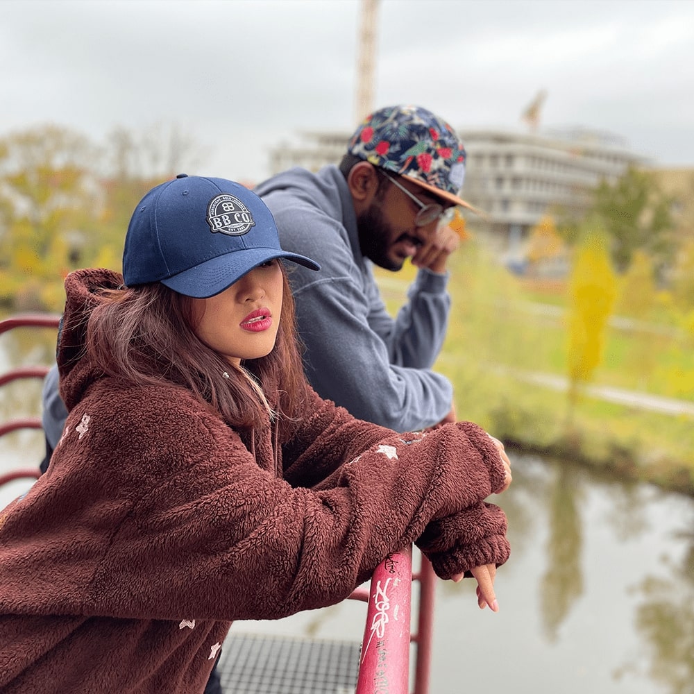 Eine Junge Frau (trägt blaue XXL Baseball Cap) und ein junger Mann (trägt extragroße Fivepanel Cap) lehnen an einem Geländer und schauen auf einen Fluss.