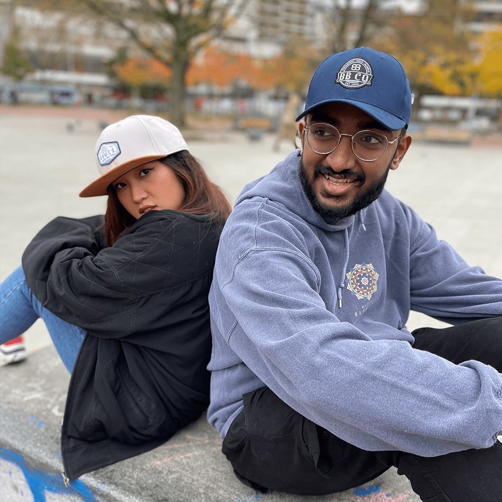 Eine junge Frau und ein junger Mann sitzen Rücken an Rücken auf einer Bank. Der junge Mann trägt die extragroße, blaue Baseball Cap. Die junge Frau trängt die graue XXL Snapback Cap von BigBrains Co.