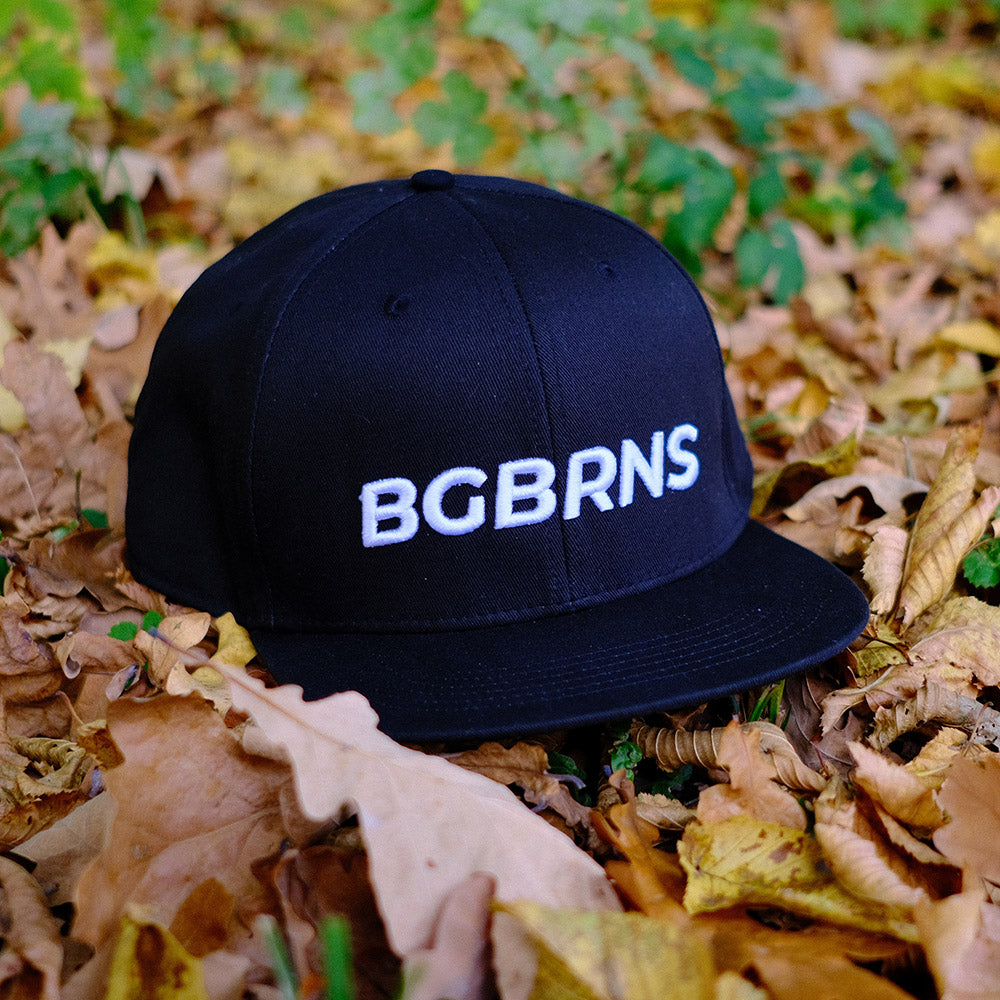 Die schwarze oversize Snapback liegt im herbstlichen Laub. Mit einer weißen Aufstickung ist der Schriftzug "BGBRNS" auf der Cap.