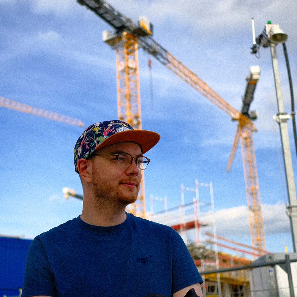 Niklas Hermsdorf trägt die bunte oversize Fivepanel Cap von BigBrains Co. Im Hintergrund sind mehrere Kräne.