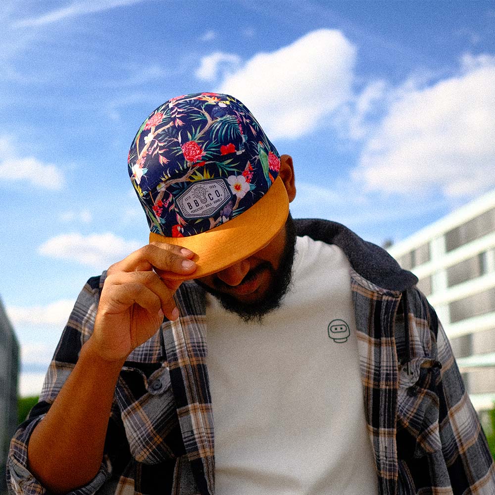 Junger Mann hält sich mit einer Hand an die übergroße, bunte Fivepanel Cap von BigBrains Co. in der XL Größe für große Köpfe.