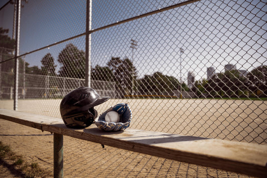 Ein Baseball Helm, sowie ein Handschuh liegen auf einer Bank. Im Hintergrund ist ein Zaun, der das Baseball Spielfeld abgrenzt.