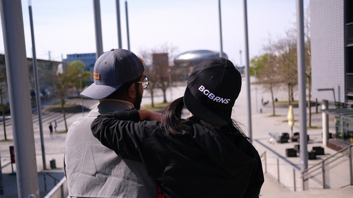 Eine junge Frau und ein Mann stehen Schulter an Schulter, weggedreht von der Kamera. Sie tragen die extragroße Trucker Cap und schwarze Snapback Cap von BigBrains Co.