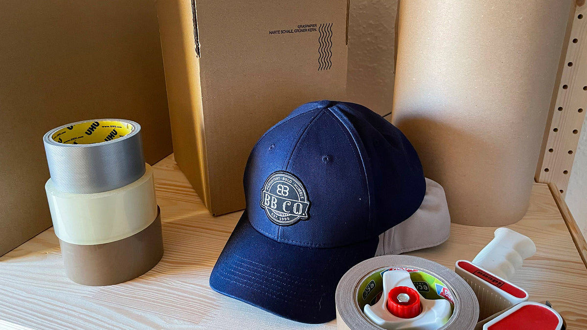 Regalplatz mit einer blauen XXL Baseball Cap von BigBrains Co., Paketklebebändern, einem Abroller und nachhaltigen Graspapierkartons.