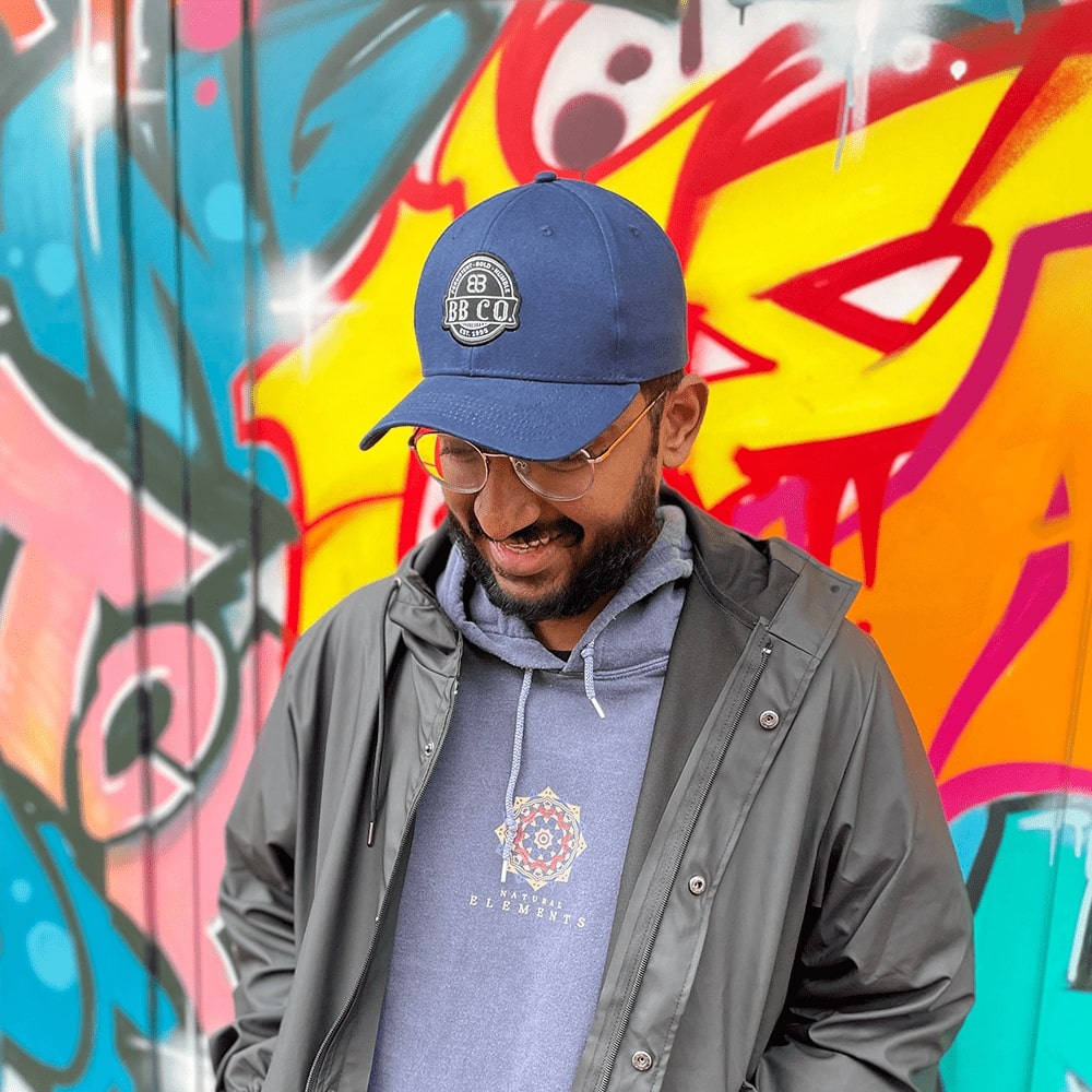Junger Mann der eine BigBrains Co. blaue, extragroße Baseball Cap trägt und vor einer Grafitti-Wand steht.