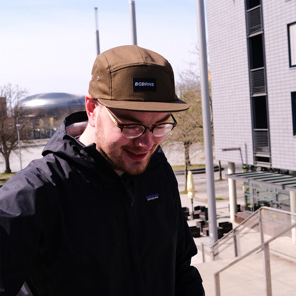 Ein Mann mit Brille trägt die dungelgrüne Fivepanel Cap von BigBrains Co. Im Hintergrund ist das Messegelände Hannover Laatzen zu sehen.