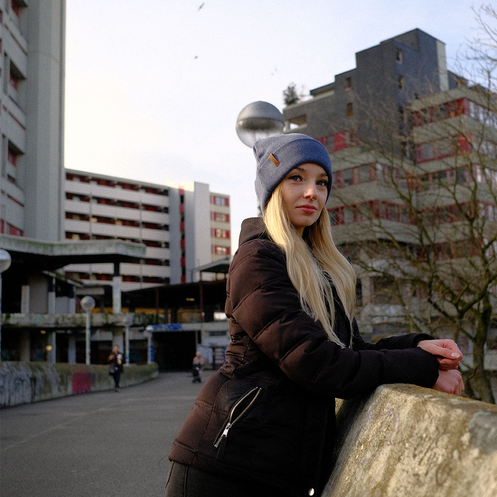Junge, blonde Frau lehnt sich am Geländer einer Brücke an. Im Hintergrund sind Hochhäuser und Plattenbauten. Die junge, blonde Frau trägt eine extragroße Beanie Mütze für große Köpfe von BigBrains Co.